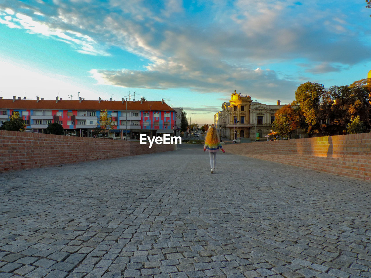 REAR VIEW OF PERSON WALKING ON FOOTPATH BY STREET AGAINST SKY