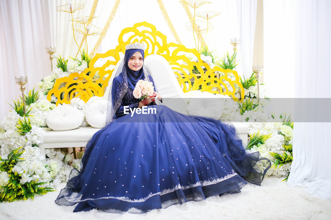 Portrait of beautiful bride holding flower bouquet while sitting on sofa 