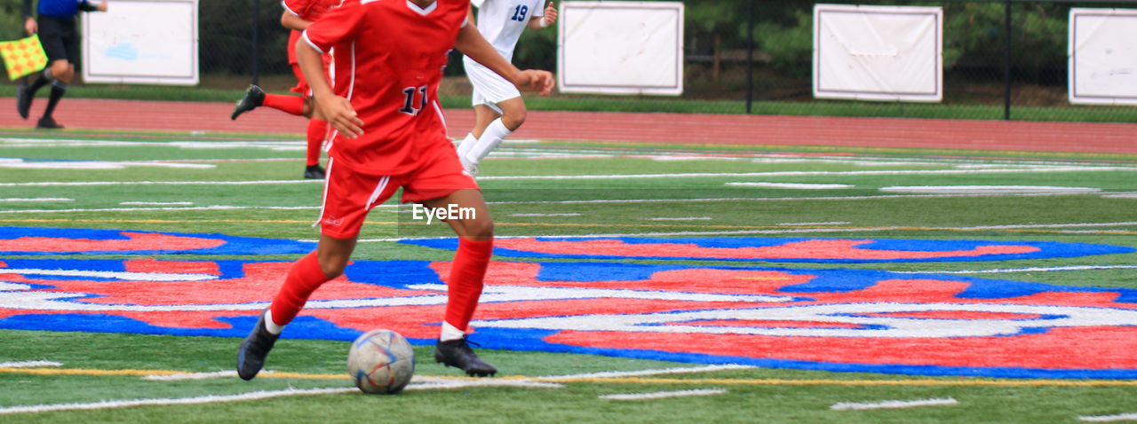 Front view of a high school boy dribbling the soccer ball up field during a game on a turf field.