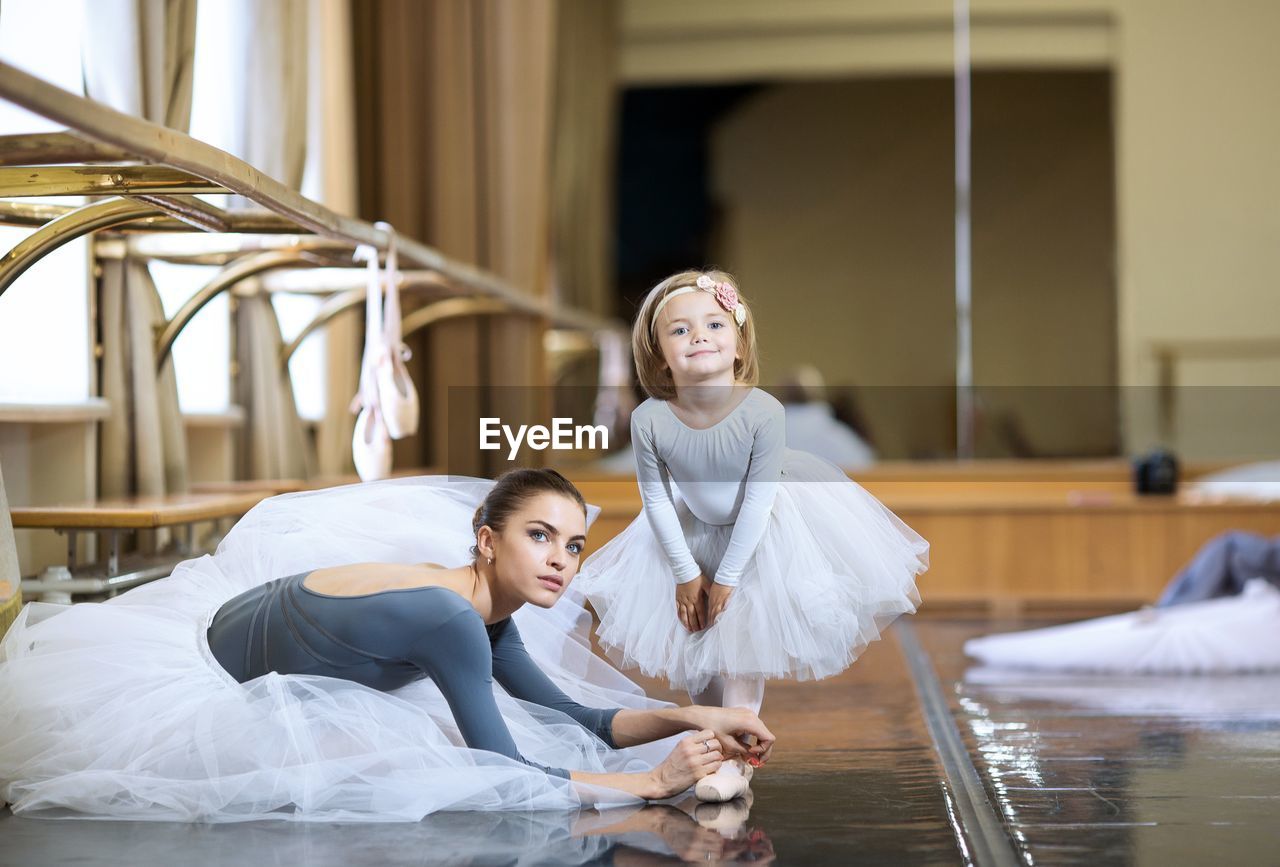 An adult ballerina is tying pointe shoes to a little ballerina.