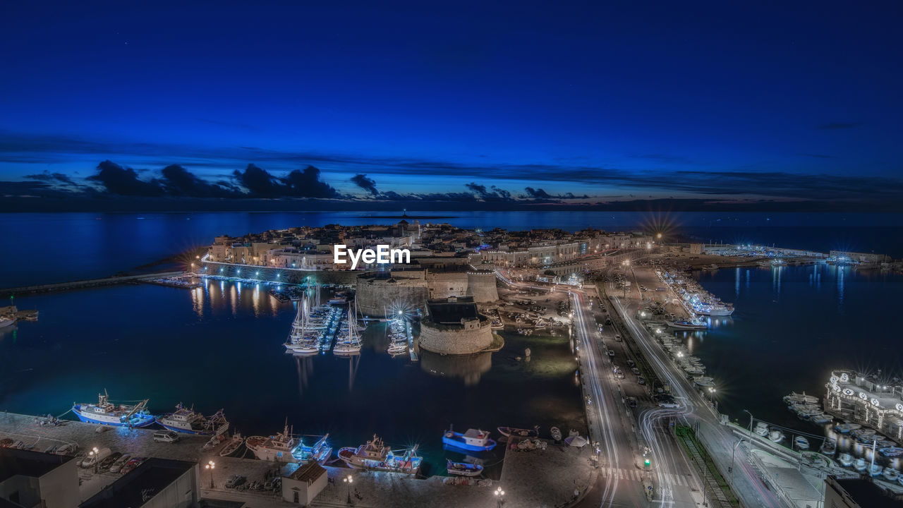 HIGH ANGLE VIEW OF ILLUMINATED CITY BY SEA AGAINST SKY