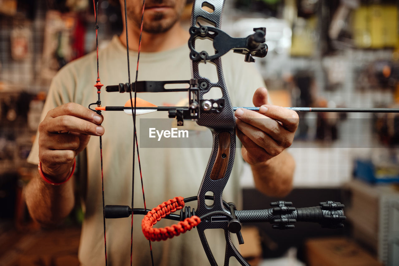 Cropped unrecognizable busy male hunter adjusting compound bow with arrow while standing in garage and preparing for shooting