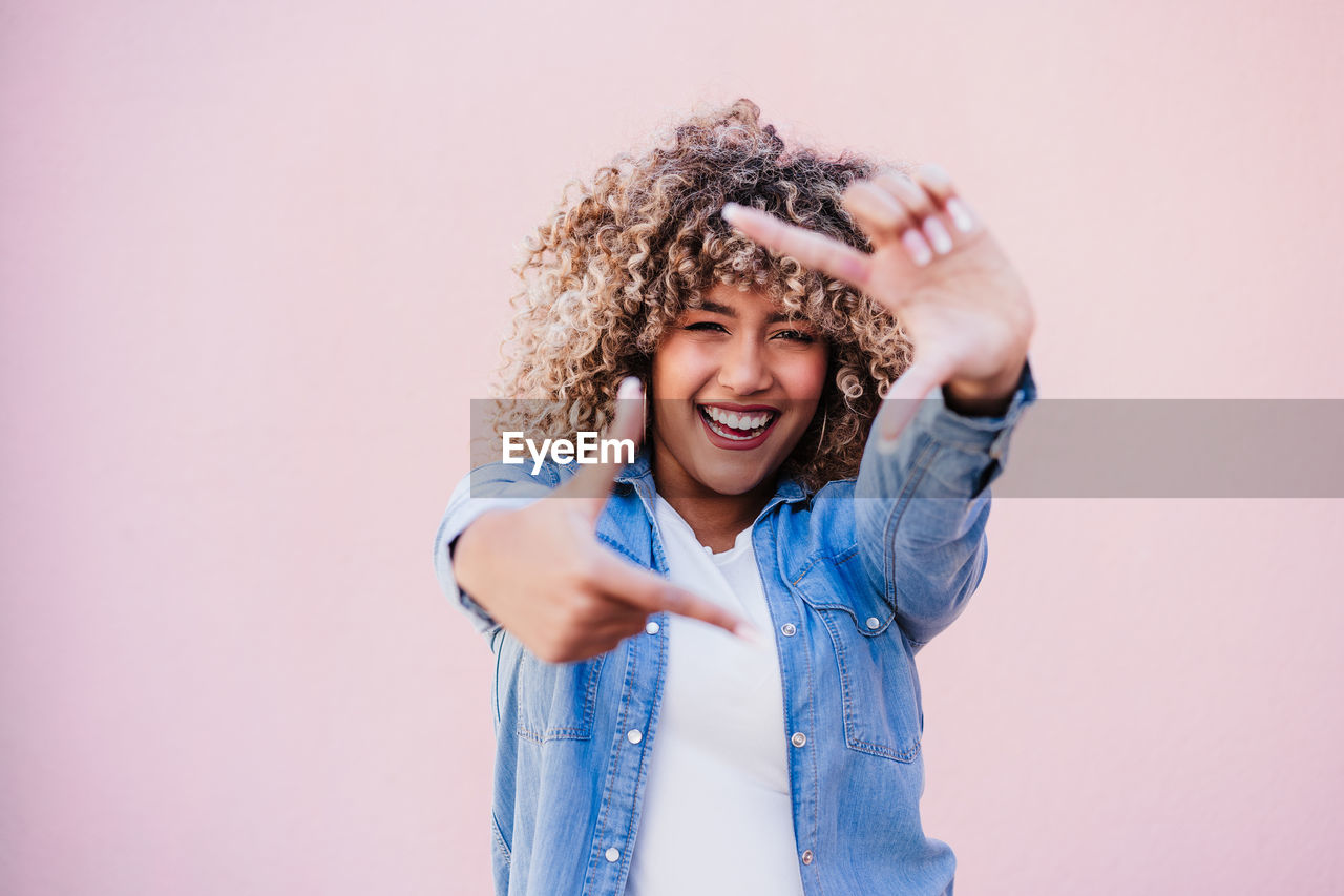 Portrait of beautiful hispanic woman afro hair outdoors in spring doing frame with hands. pink wall