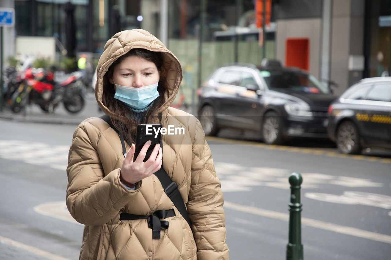 A young woman in a warm down jacket in a hood and in a mask reads a message