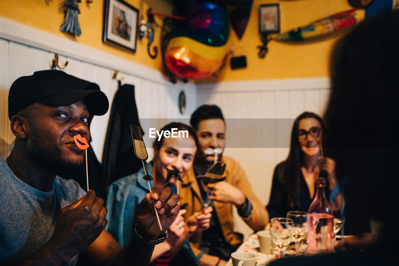 Young friends holding props while sitting at restaurant during dinner party