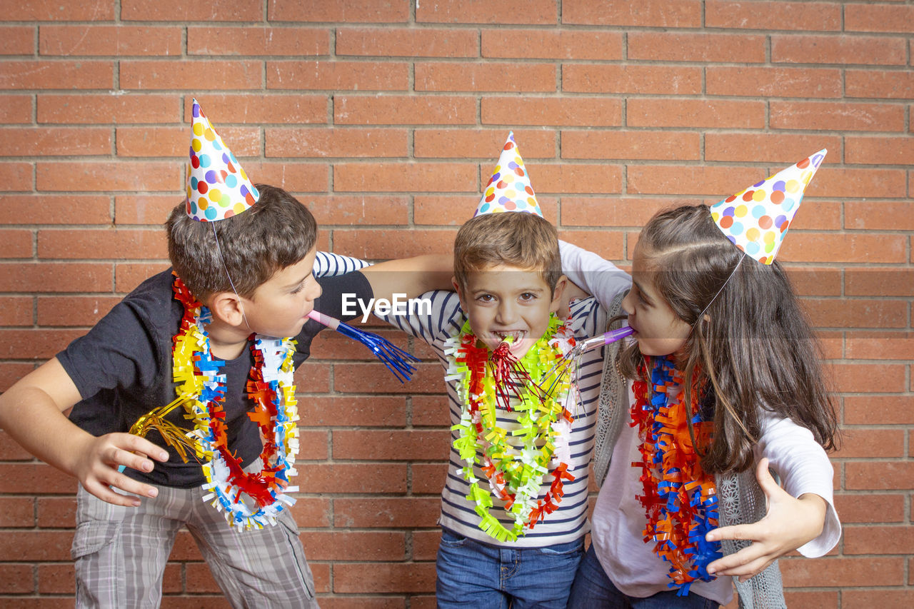 Portrait of siblings wearing garlands and party hats against brick wall