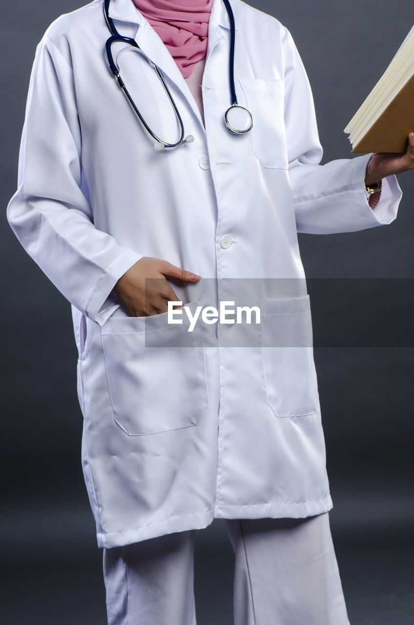 Midsection of doctor reading book while standing against gray background