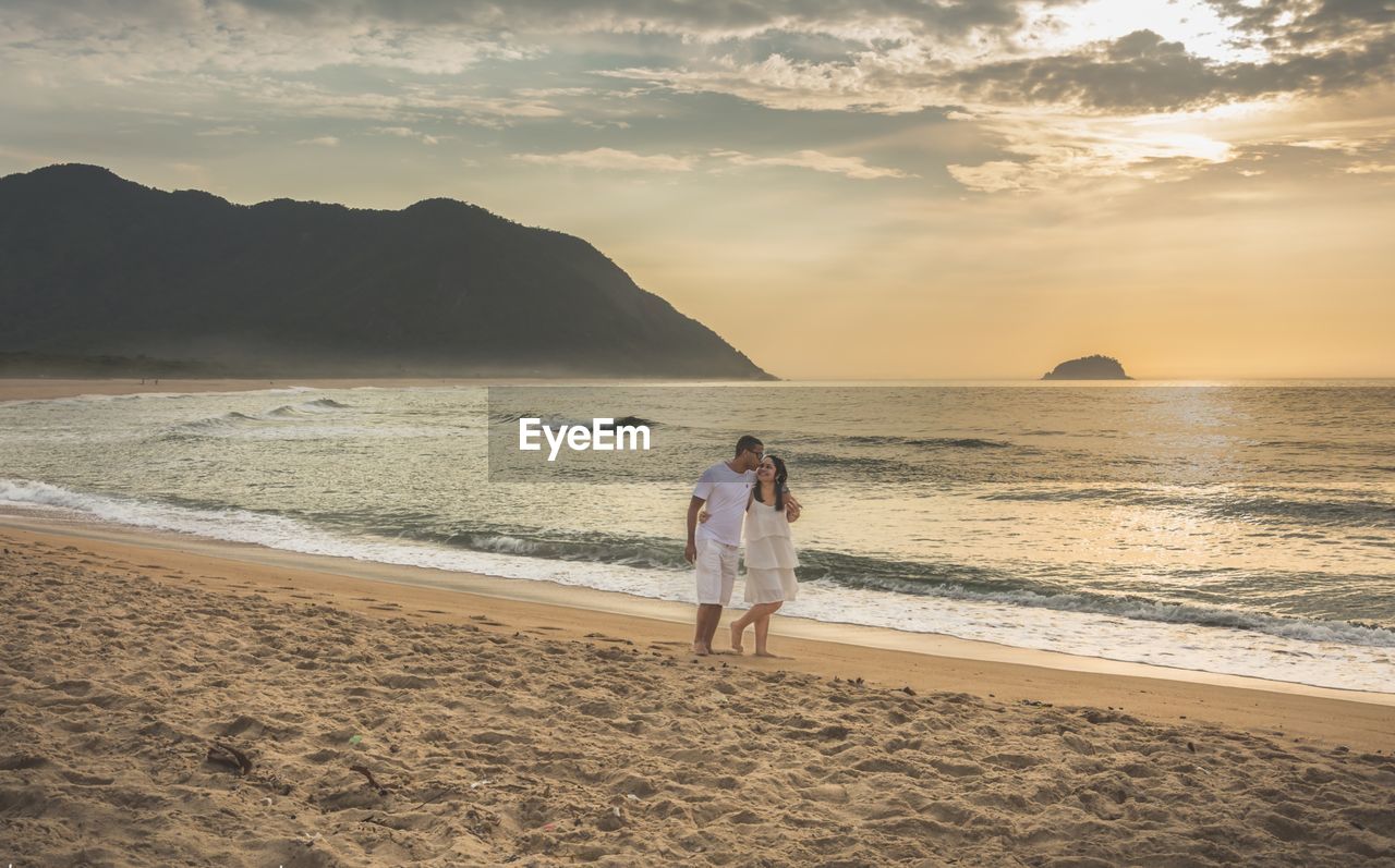Smiling couple walking at beach against sky during sunset
