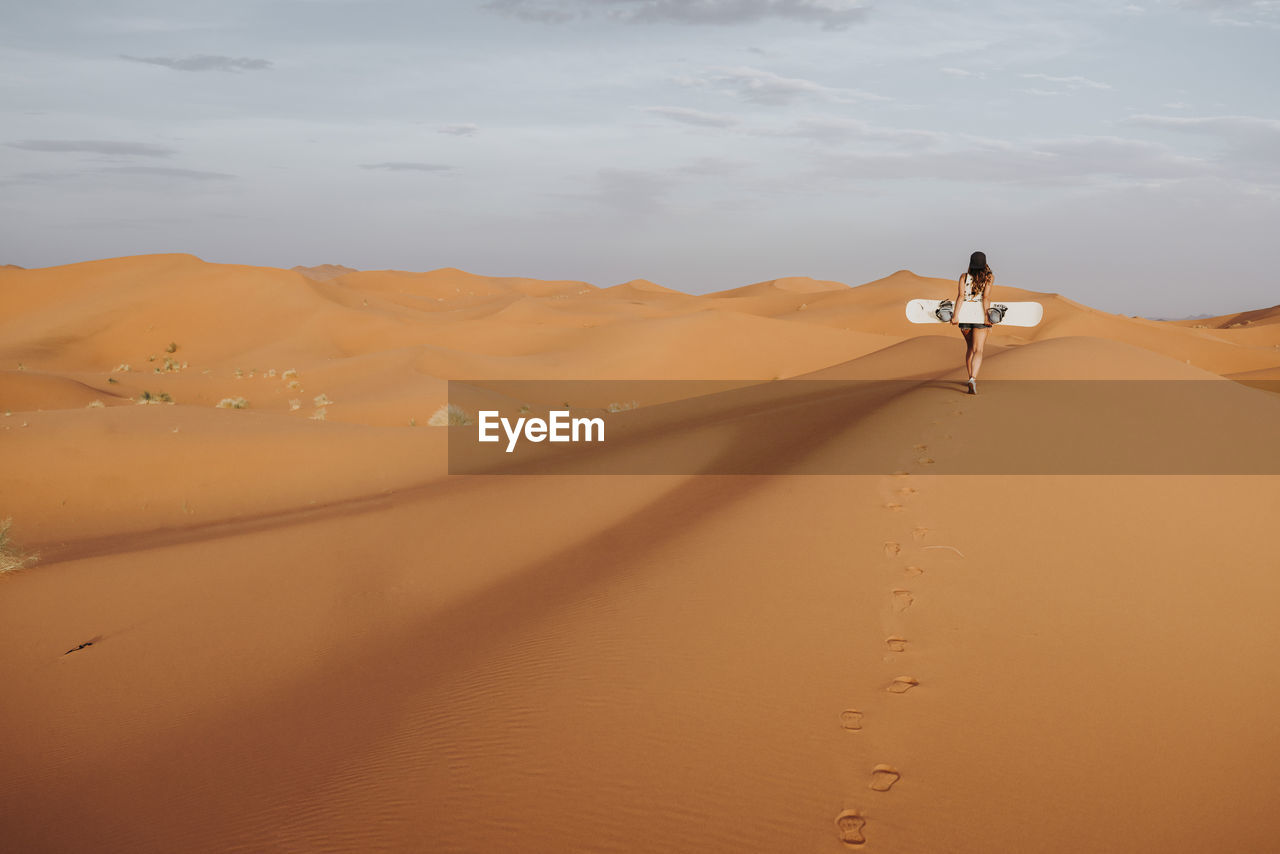 Back view of young female walking on sand and preparing for sandboarding looking away