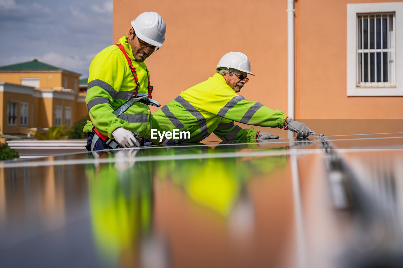 Group of workers in uniform and helmets installing photovoltaic panels on roof of wooden construction near house