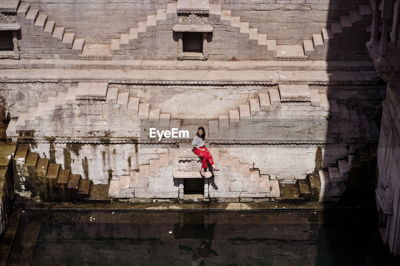 From above young ethnic female tourist sitting on steps of toorji well with water during sightseeing in india