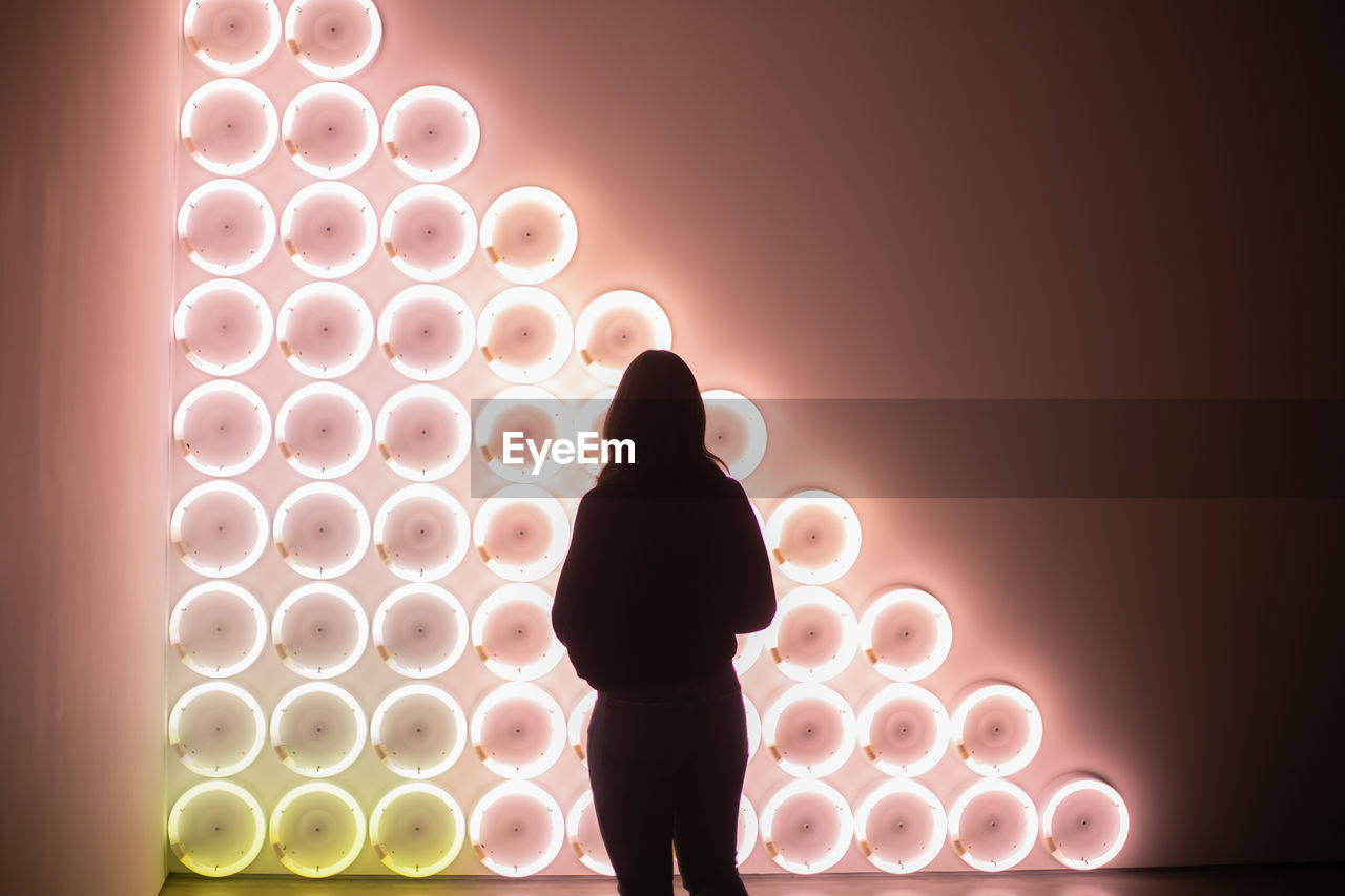 REAR VIEW OF SILHOUETTE WOMAN STANDING AGAINST ILLUMINATED LIGHT