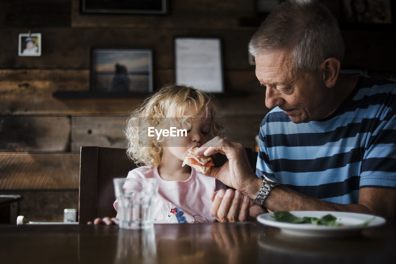 Grandfather feeding food to granddaughter while sitting at table in home