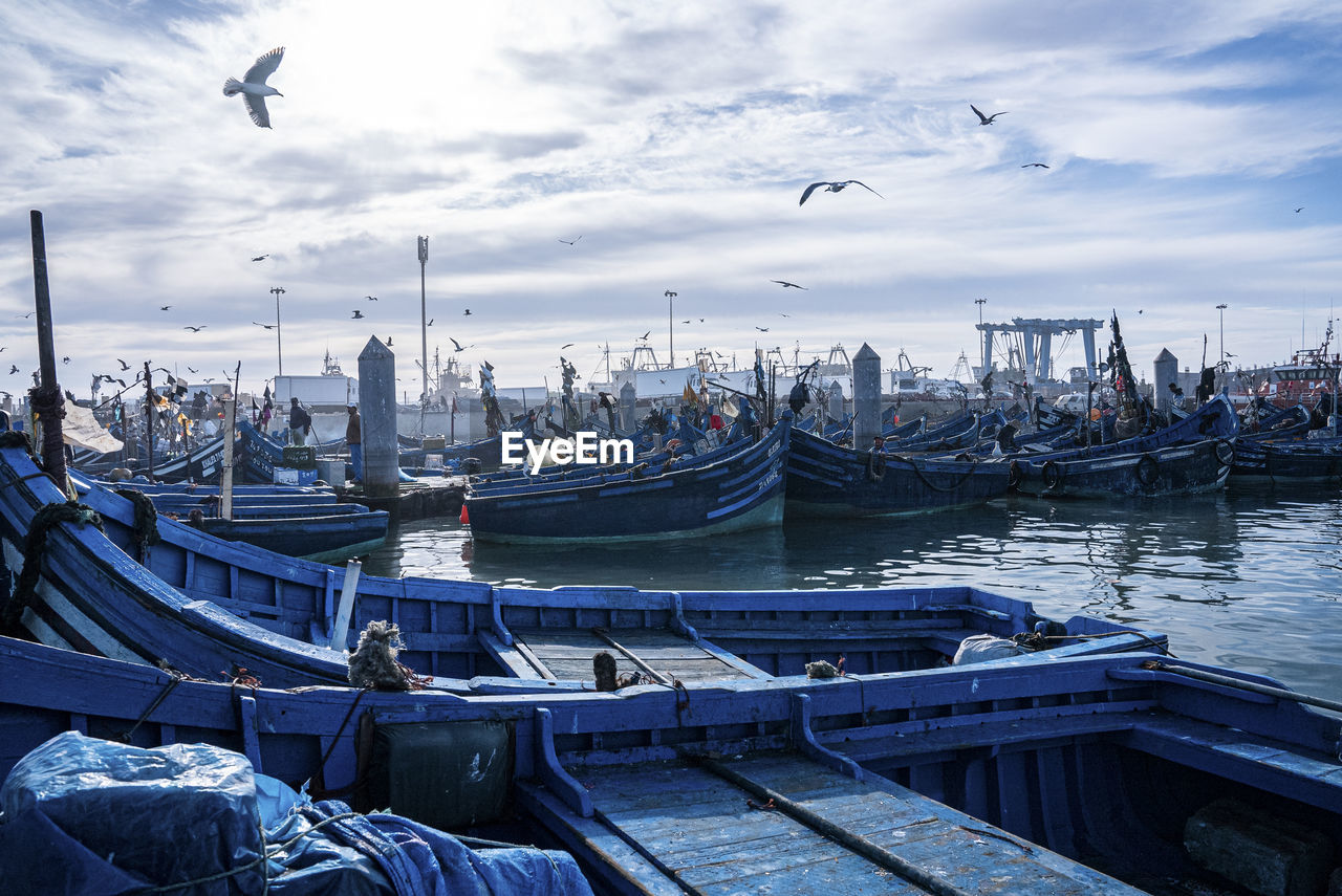 Wooden blue fishing boats anchored at marina against cloudy sky