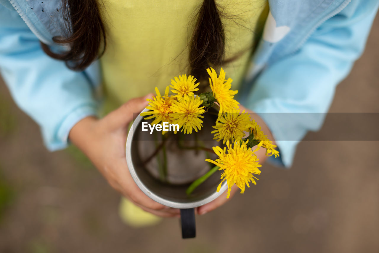 A girl in a blue jacket holds a bouquet of dandelions in an iron mug.