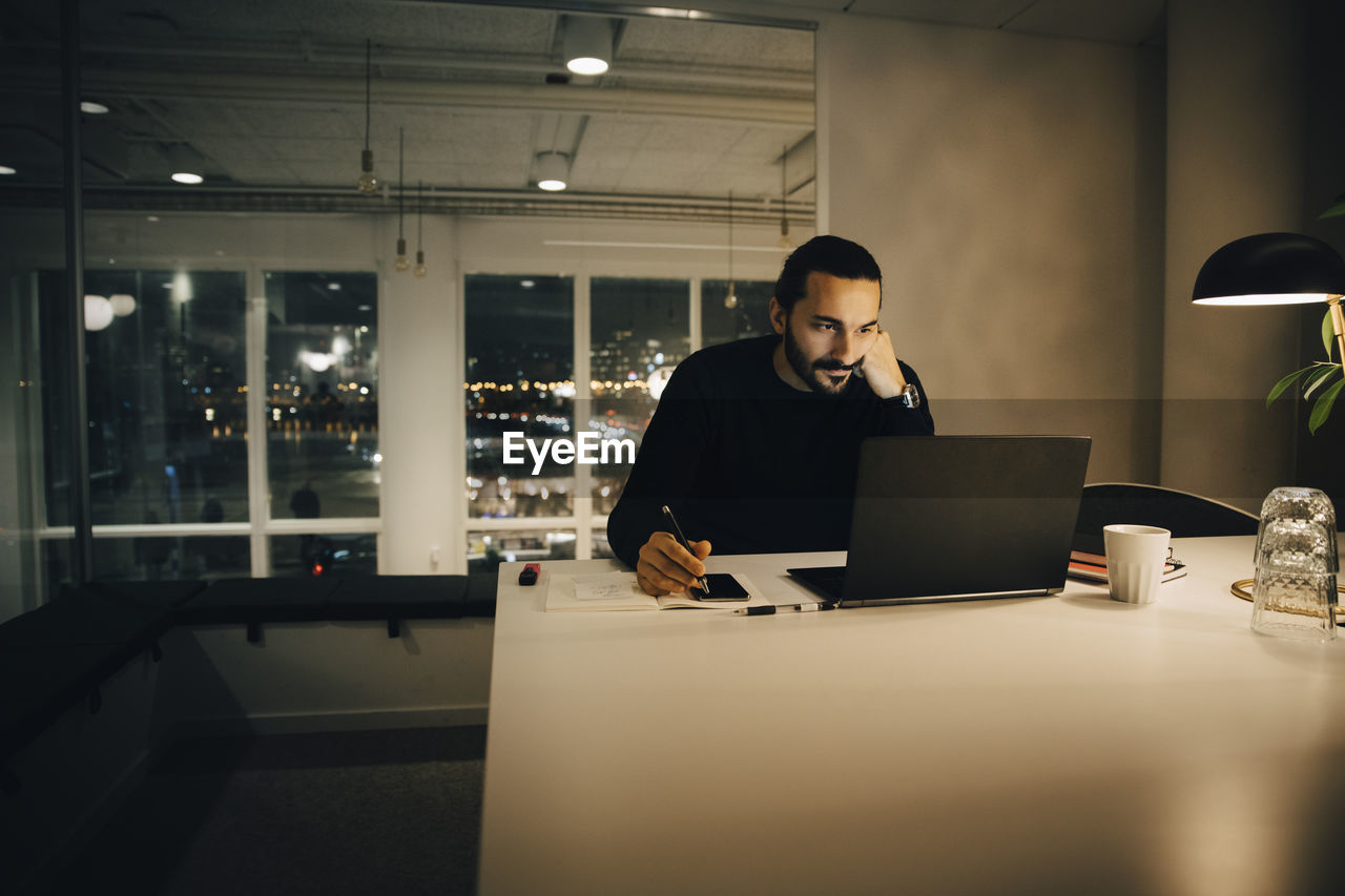 Tired businessman working late while sitting at desk in illuminated creative office