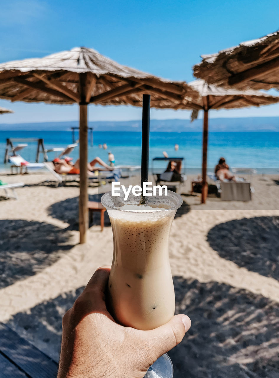 Personal perspective of person holding glass of cold iced coffee on beach in summer.