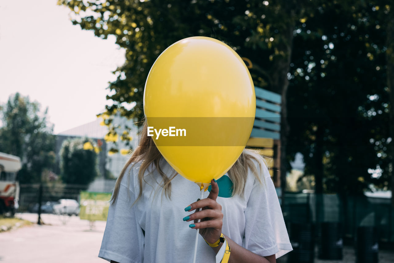 Woman holding yellow balloon in front of face against trees