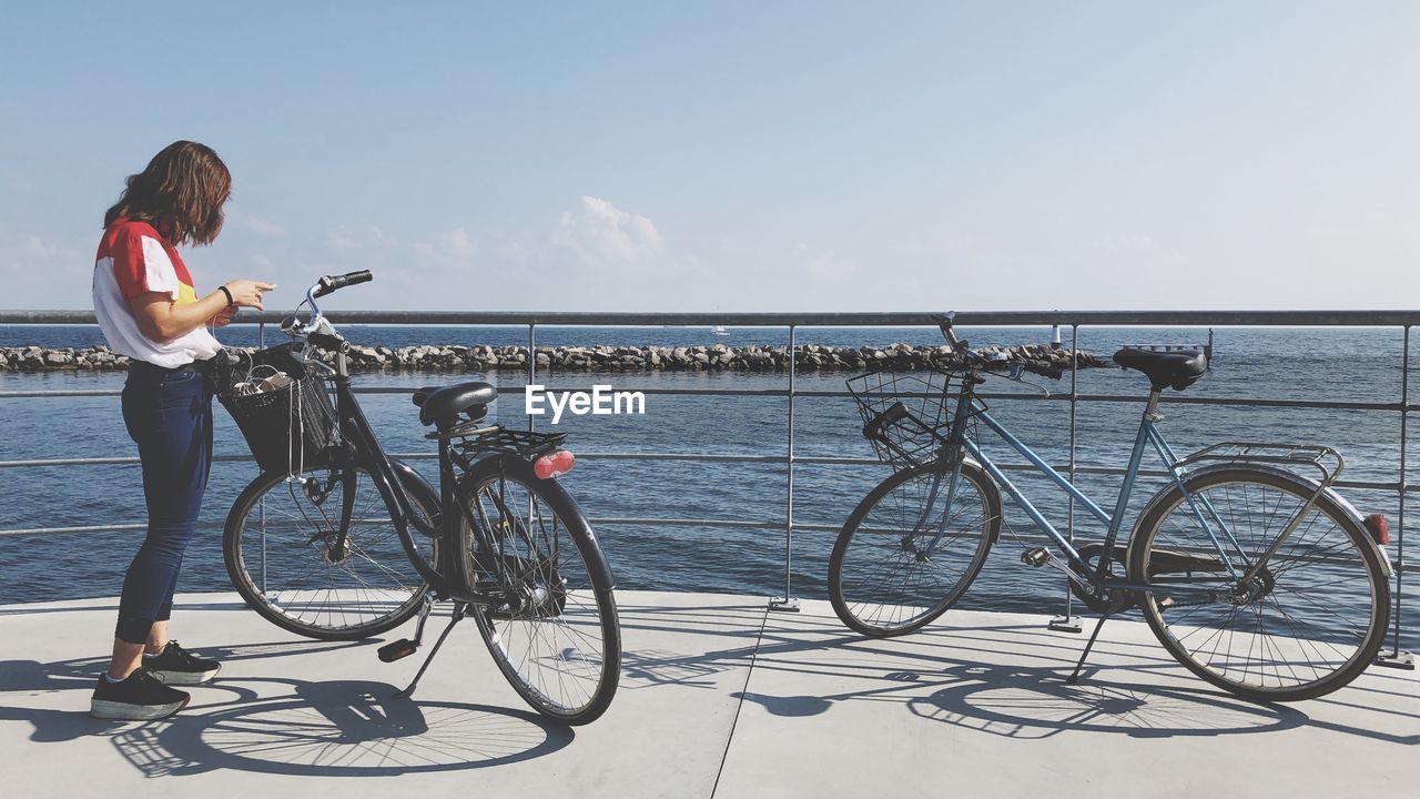 Side view of woman standing with bicycles on promenade against sky