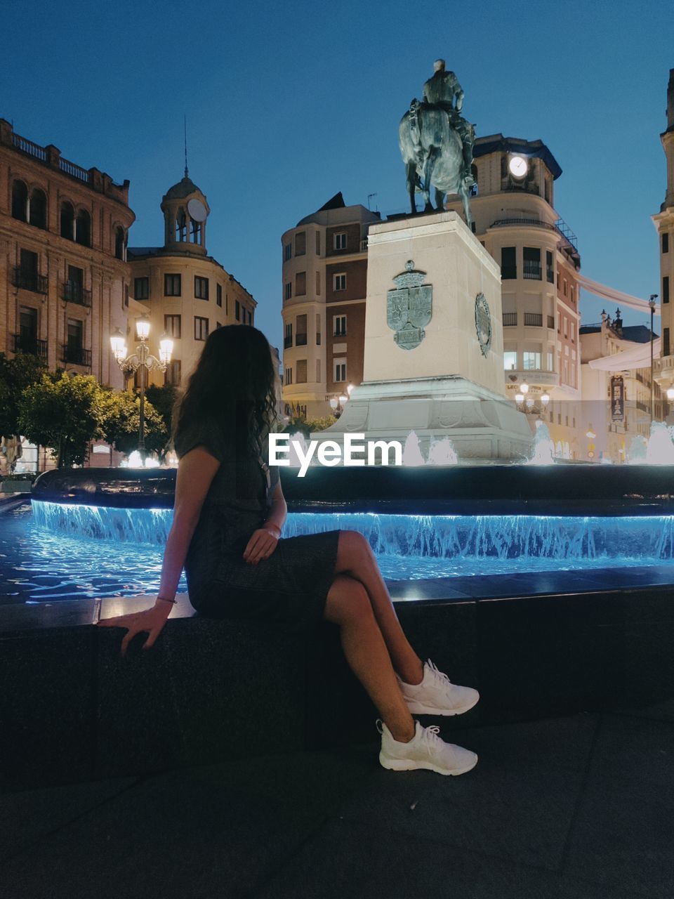 WOMAN SITTING BY SWIMMING POOL AGAINST BUILDINGS