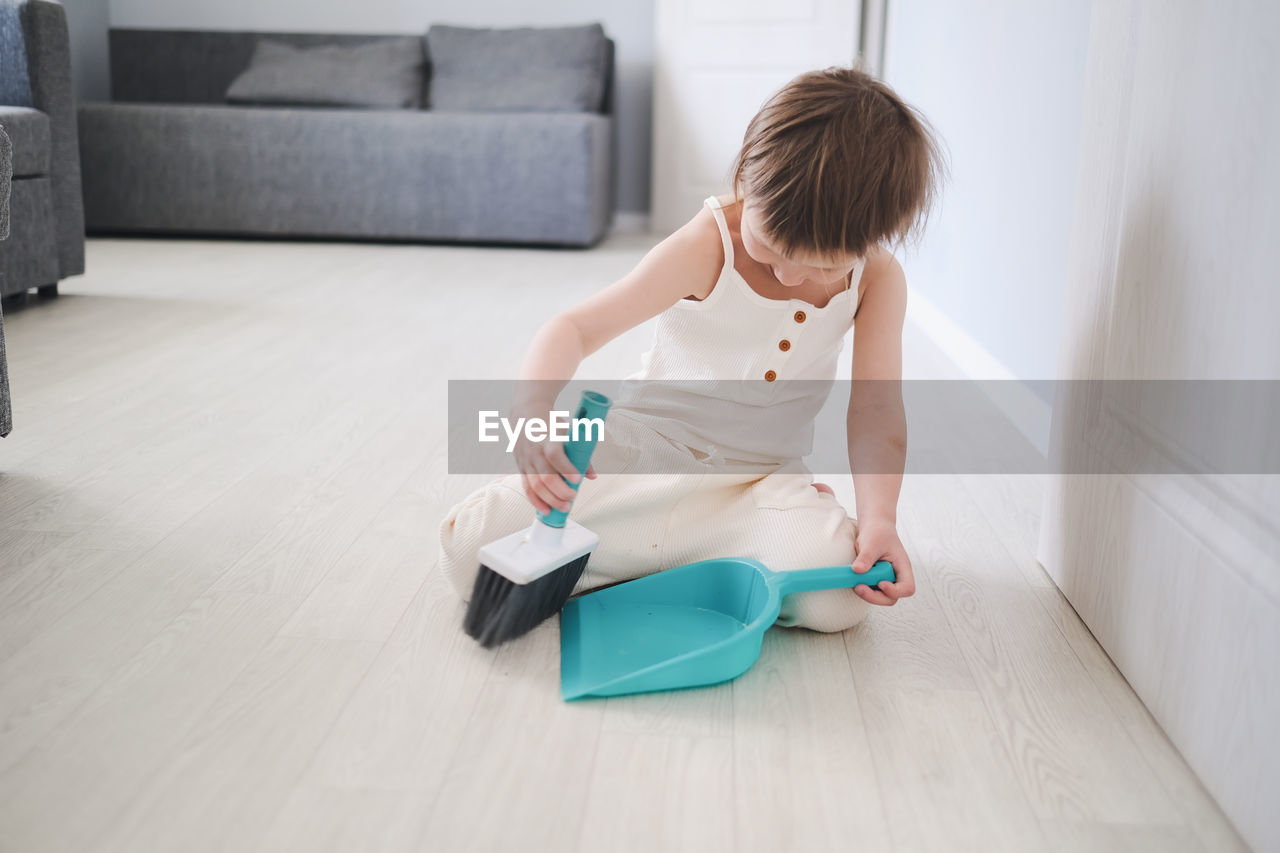 one person, indoors, sitting, flooring, domestic life, childhood, home interior, toddler, full length, child, lifestyles, living room, domestic room, hardwood floor, cleanliness, casual clothing, floor, furniture, person, looking down, wood, looking, women, hairstyle, clothing, adult, holding, day, concentration, kneeling