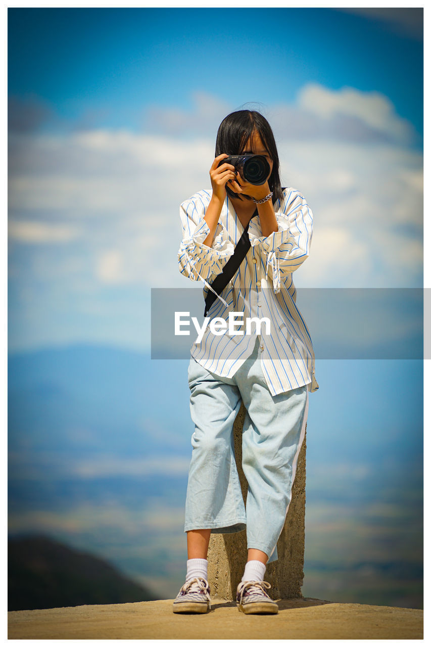 Low angle view of girl photographing while standing on rock against sky