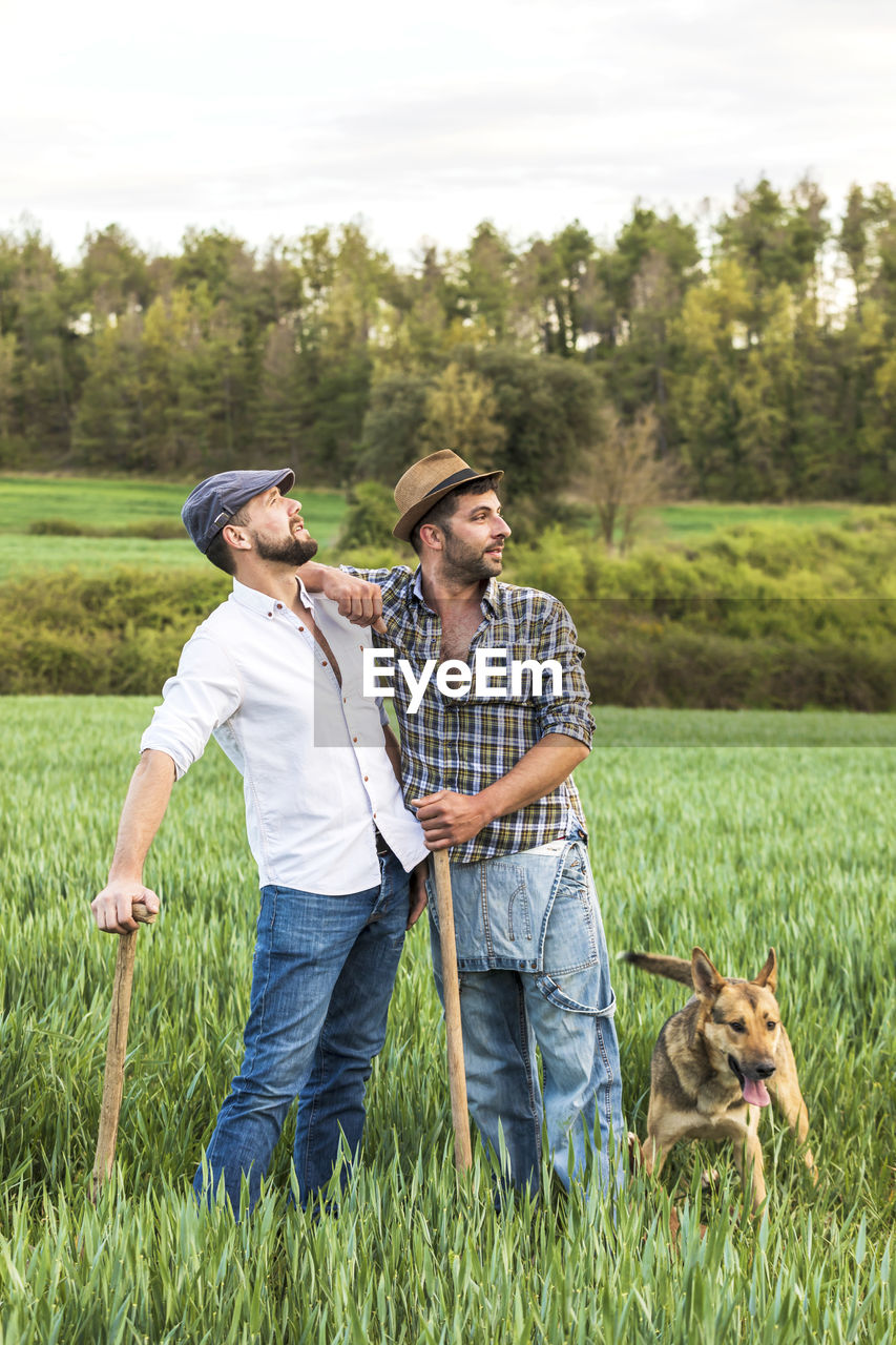 Male farm workers with dog standing on agricultural field against sky during sunset