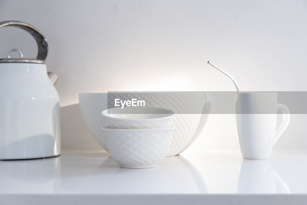 White ceramic bowls, coffee cup with metal straw and kettle set on quartz countertop in sunlight