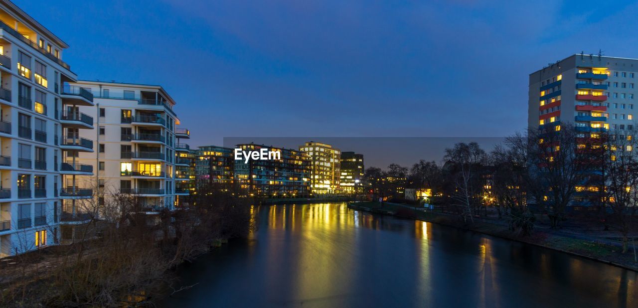 RIVER AMIDST ILLUMINATED BUILDINGS AGAINST SKY AT DUSK