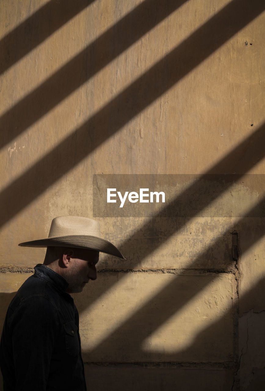 Side view of adult man in cowboy hat against wall with sunlight and shadow