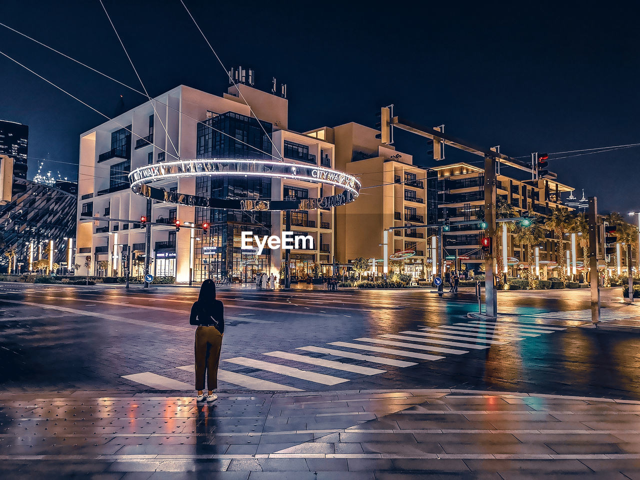 REAR VIEW OF MAN WALKING ON ILLUMINATED STREET BY BUILDINGS AT NIGHT