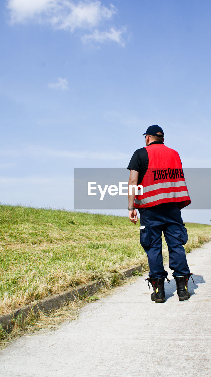 Rear view of firefighter walking on road by grass field against sky