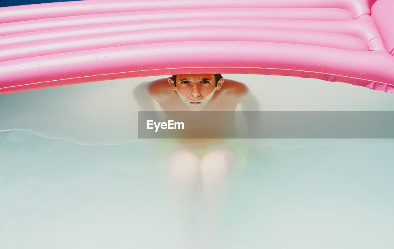 Boy in a pool taking a bath under a pink flat looking at camera.