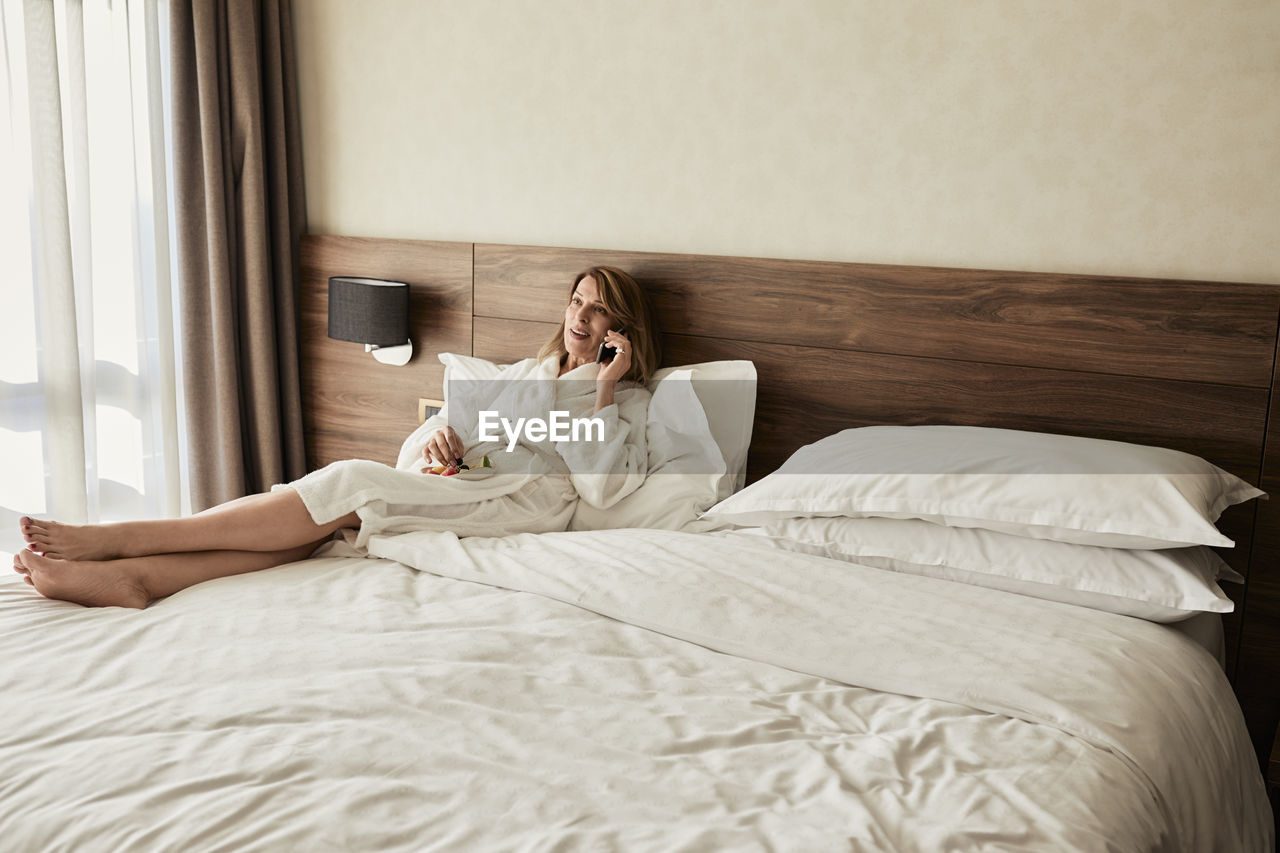 Senior woman talking through mobile phone while reclining on bed with fruit in hotel room