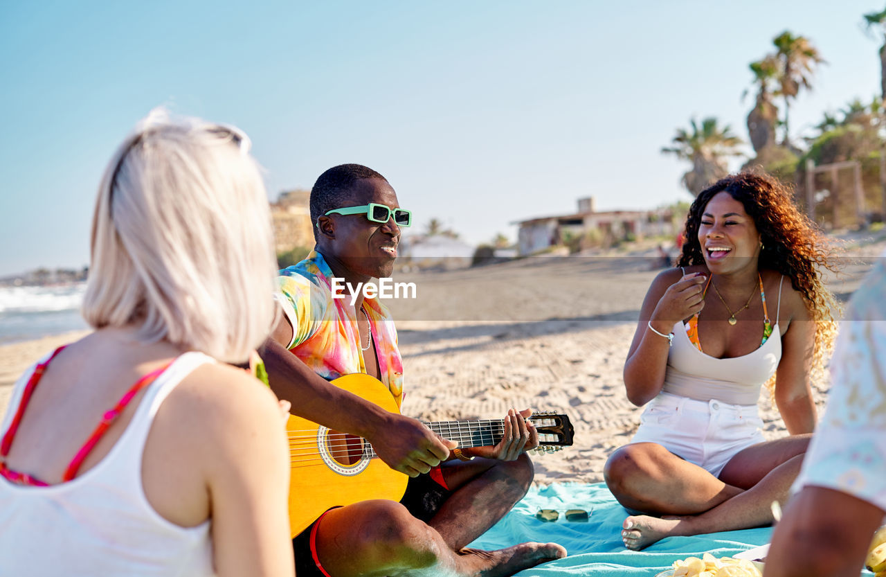 Happy multiethnic man and women in casual clothes smiling and eating fruits while sitting cross legged on blanket and listening to black friend playing acoustic guitar near sea on summer weekend day on beach