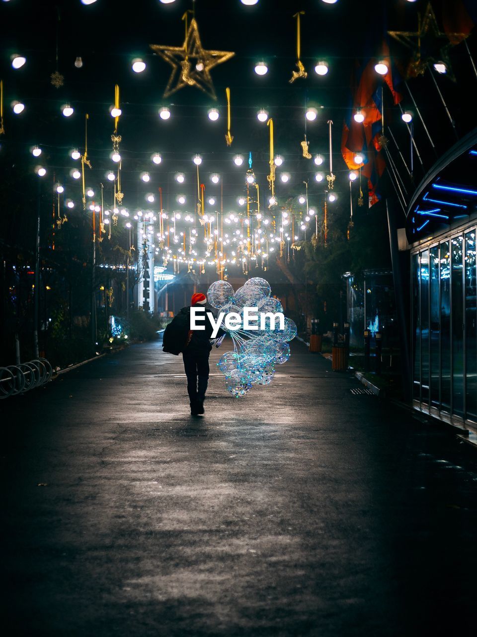 Rear view of person holding balloons while walking on illuminated street at night