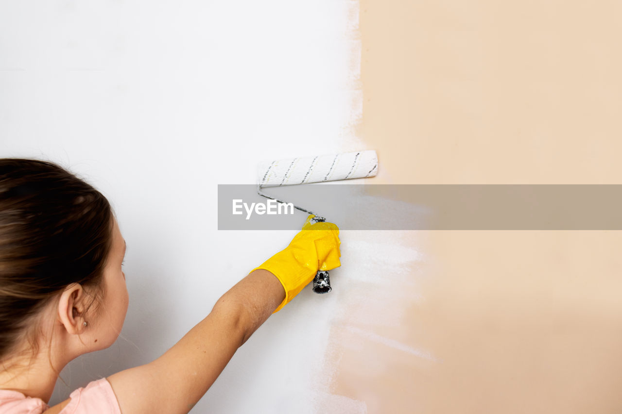 Girl child in yellow gloves paints a wall with a roller with white paint