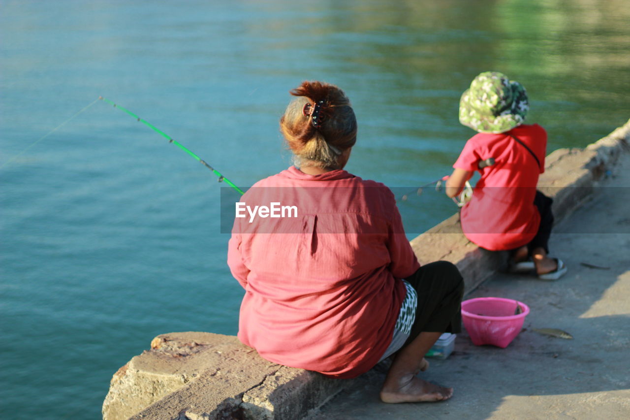 Rear view of woman and girl fishing while sitting on rock by sea