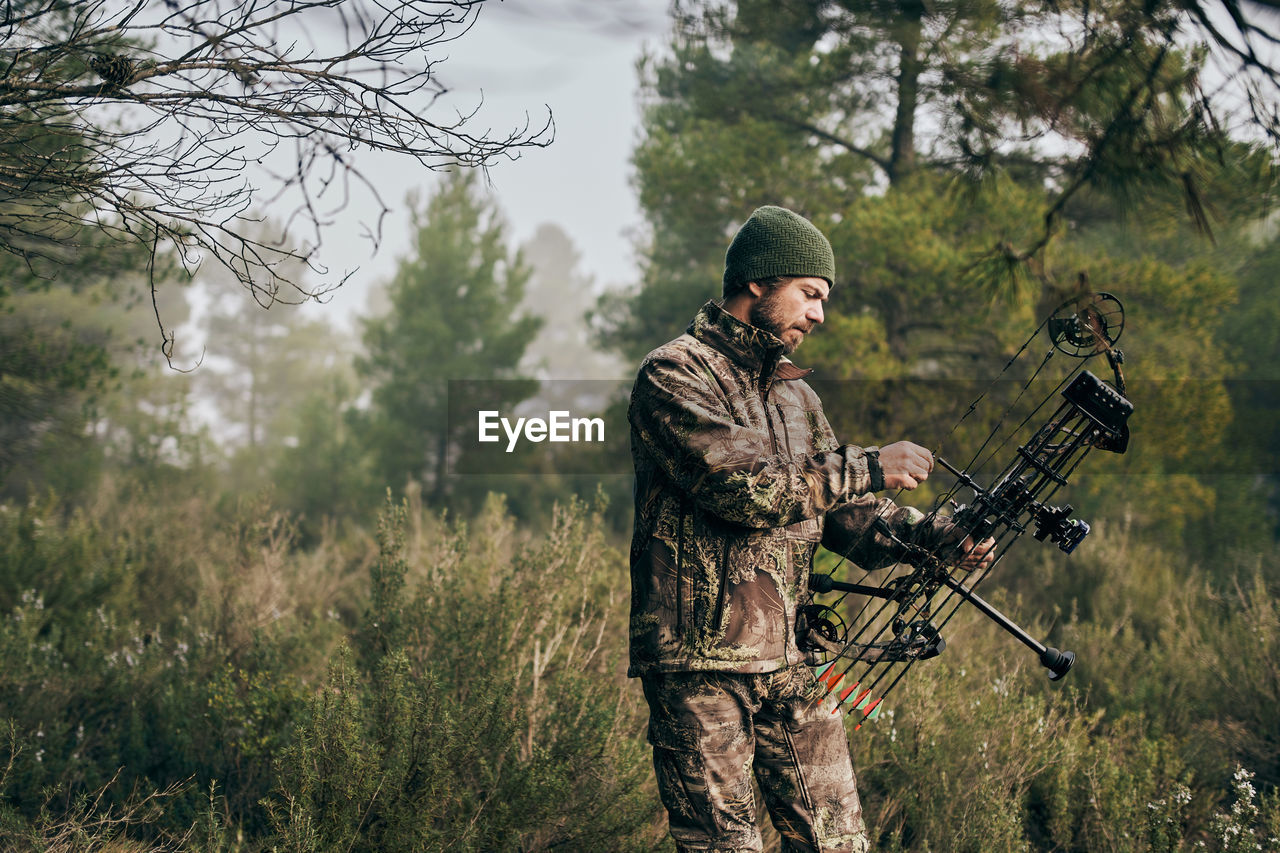 Side view of serious male hunter adjusting compound bow with arrow while preparing for hunting in woods in autumn