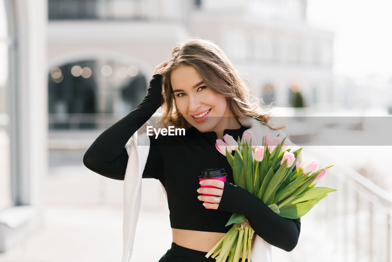 Young woman with a bouquet of spring tulips and a cup of coffee walks down the street in the city
