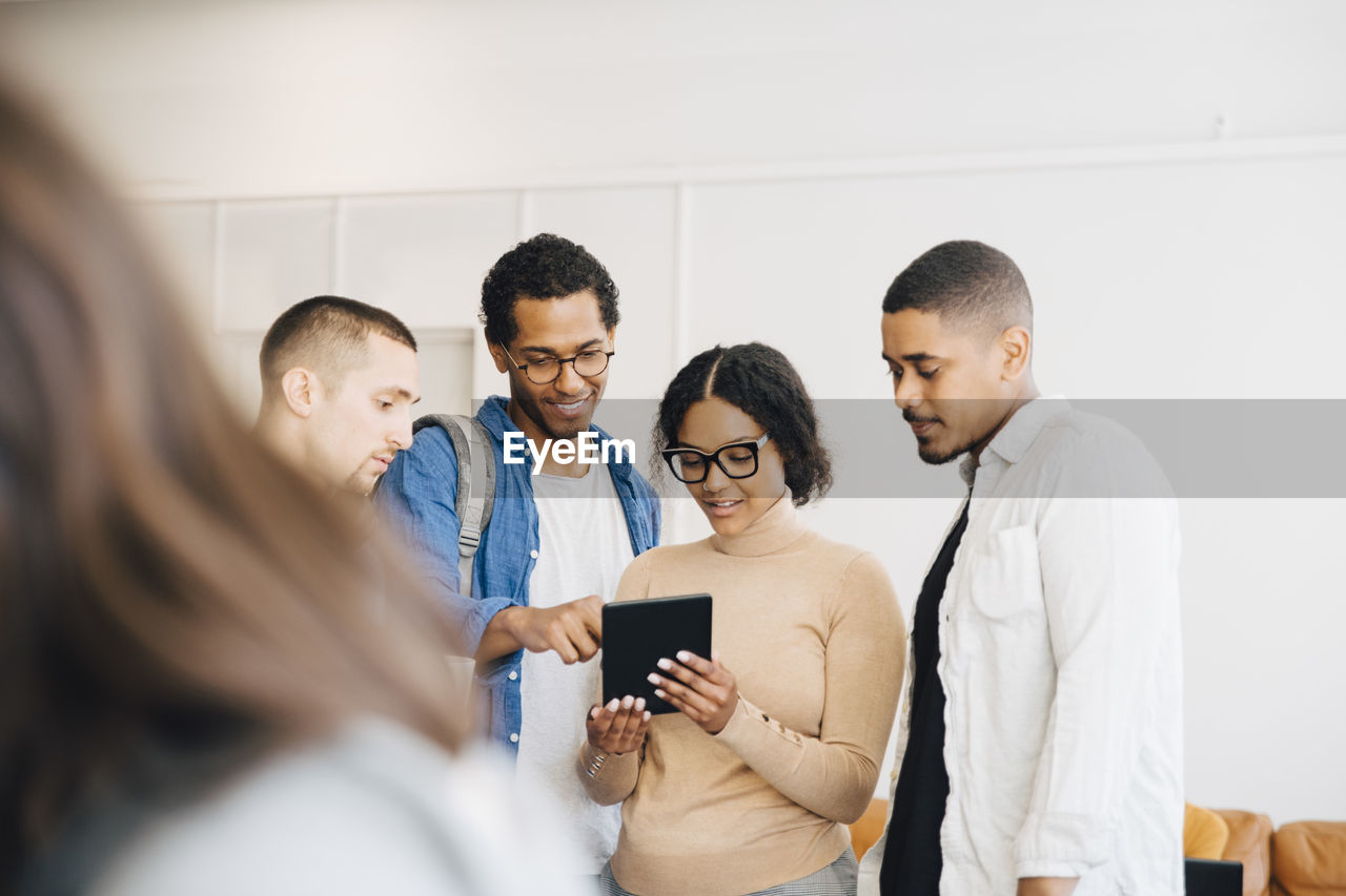 Female computer hacker showing digital tablet to male coworkers while standing against wall in office