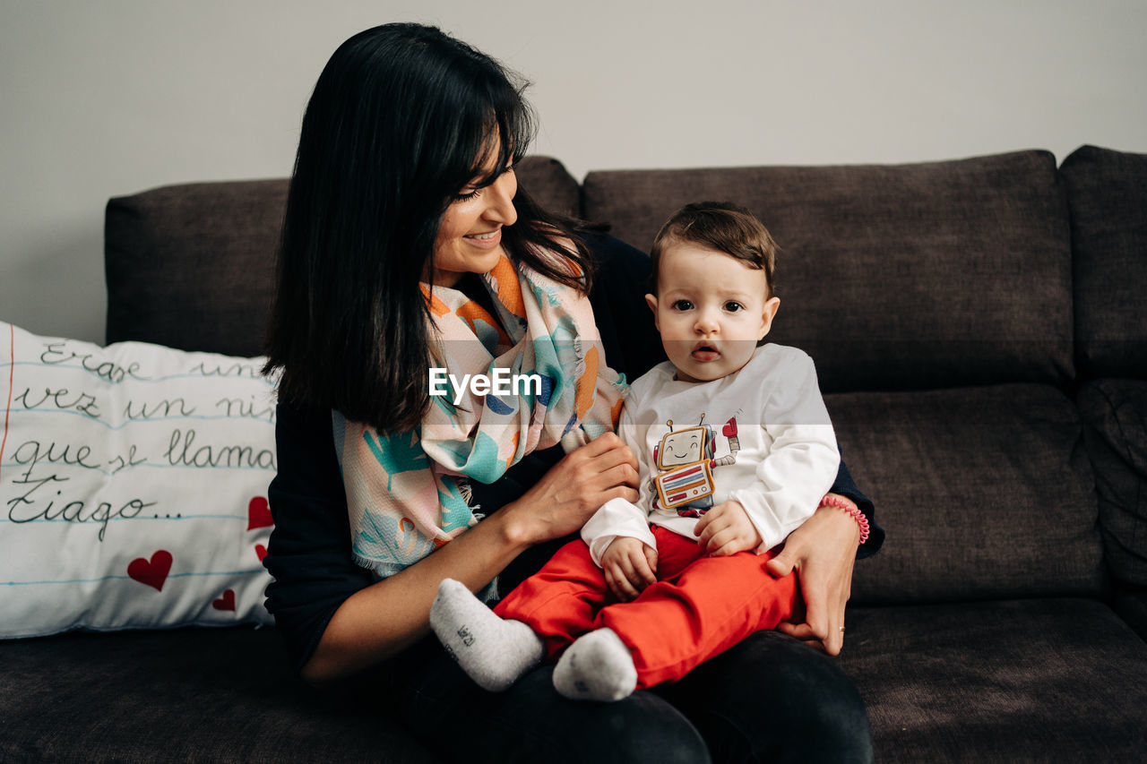 Happy ethnic woman smiling and holding cute little son on sofa at home