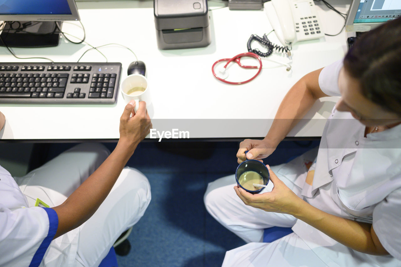 From above doctors in white uniform sitting near table in office and drinking coffee while working in hospital together
