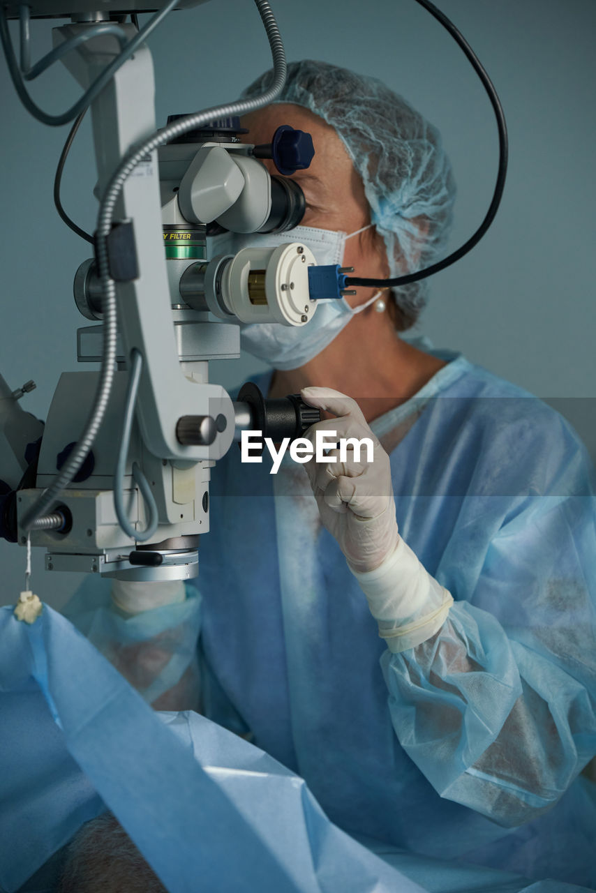 Adult female doctor in sterile mask and ornamental medical cap looking through surgical microscope against crop coworker in hospital