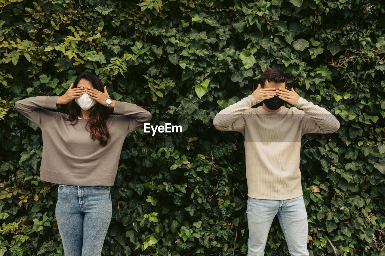 Young couple covering eyes with hands against plants in park during covid-19