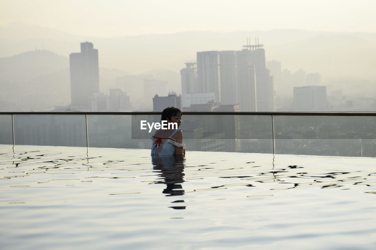 Rear view of woman in infinity pool against buildings during sunset
