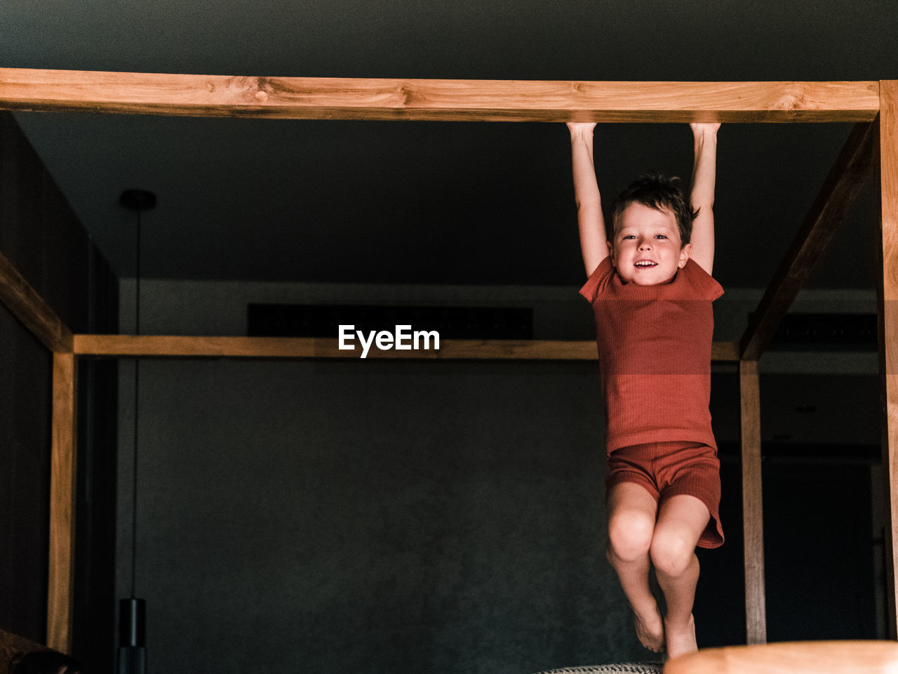 Positive little kid hanging on wooden canopy bed while having fun in bedroom and looking at camera