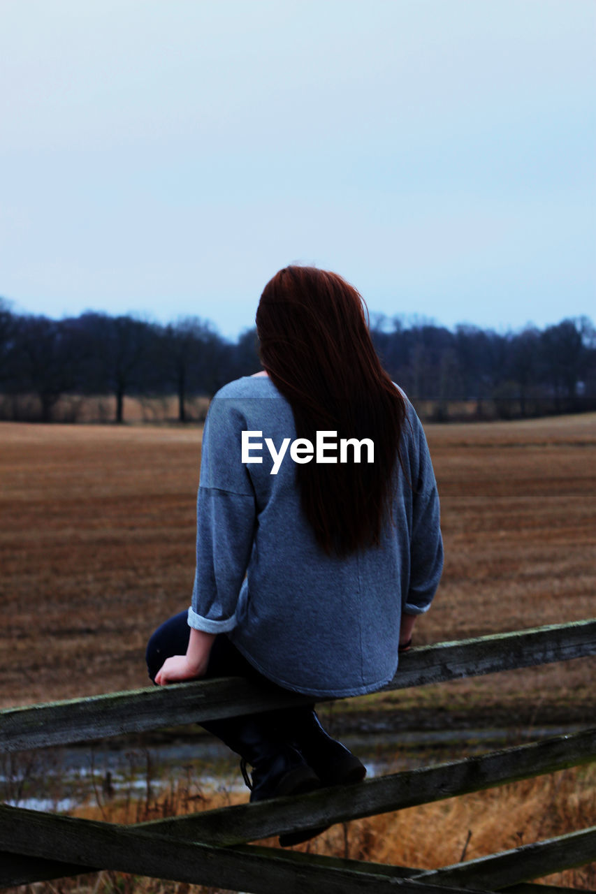 Rear view of woman sitting on railing at farm field against sky