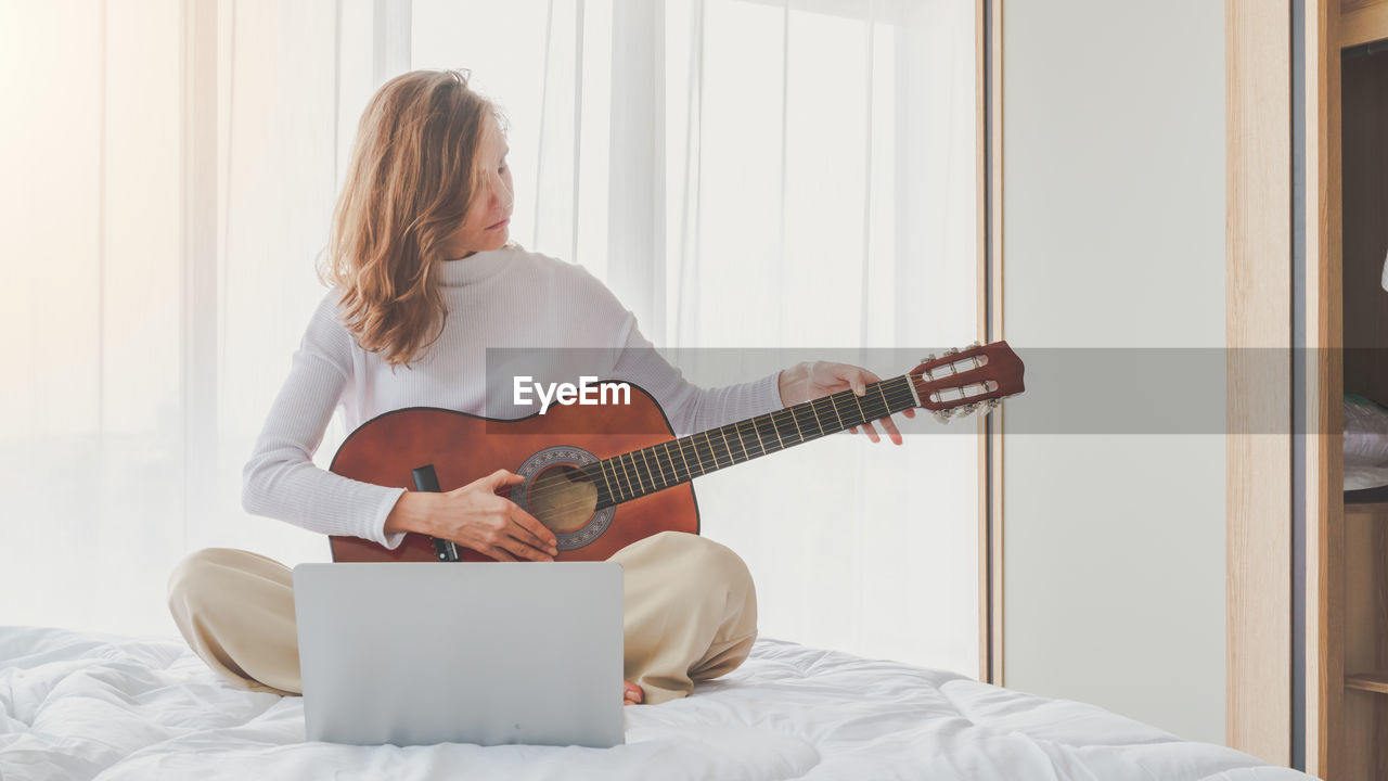 Woman playing guitar while sitting on bed at home