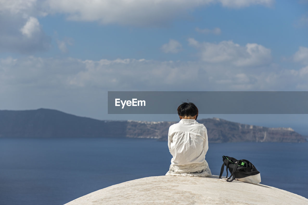 Rear view of woman sitting on rock by sea against sky